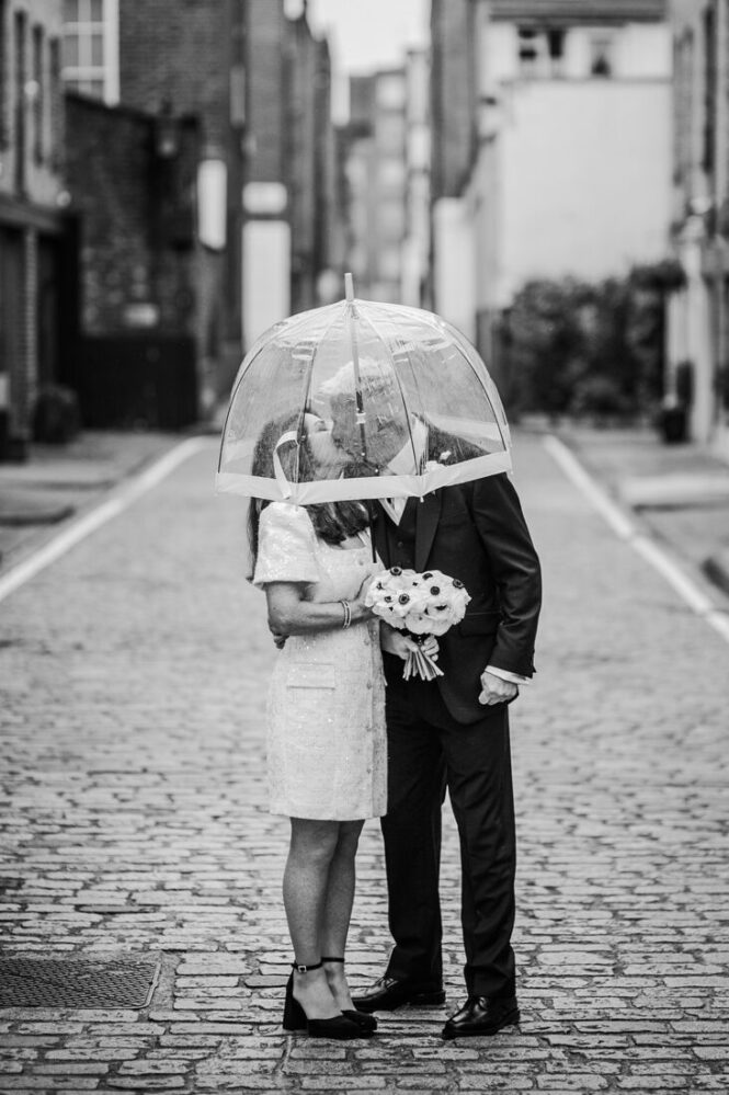 Bride and groom kiss under a transparent umbrella on a cobblestone street, bouquet in hand, in a black-and-white scene.