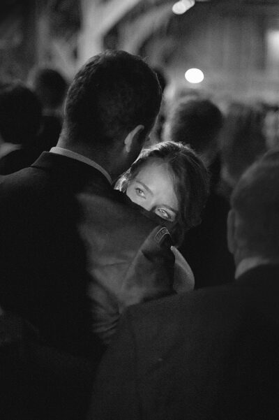 Woman peeking over a man's shoulder in a crowded, dimly lit event scene, smiling eyes visible behind him.