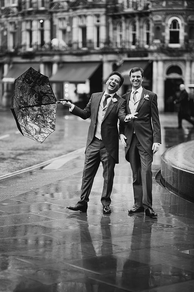 Two men in formal suits laugh on a rainy city street; one holds an umbrella over them as they pose.