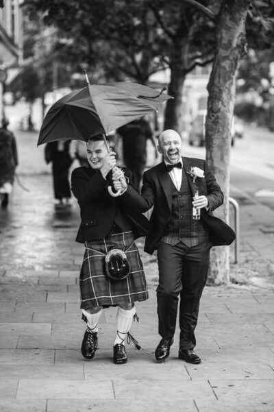 Two men in formal wear walk on a rainy city sidewalk; one holds a large umbrella while the other laughs, a bottle in hand.