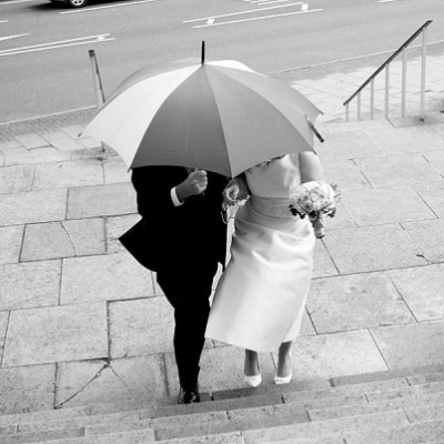 Bride and her father huddle under an umbrella as they approach the church.