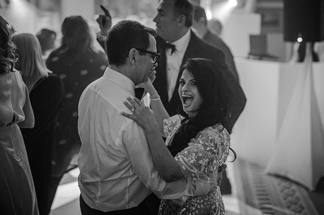 first-dance-lanesbourough-london - Wedding photographer London Black-and-white photo of a smiling couple dancing closely at a wedding reception, surrounded by other guests.