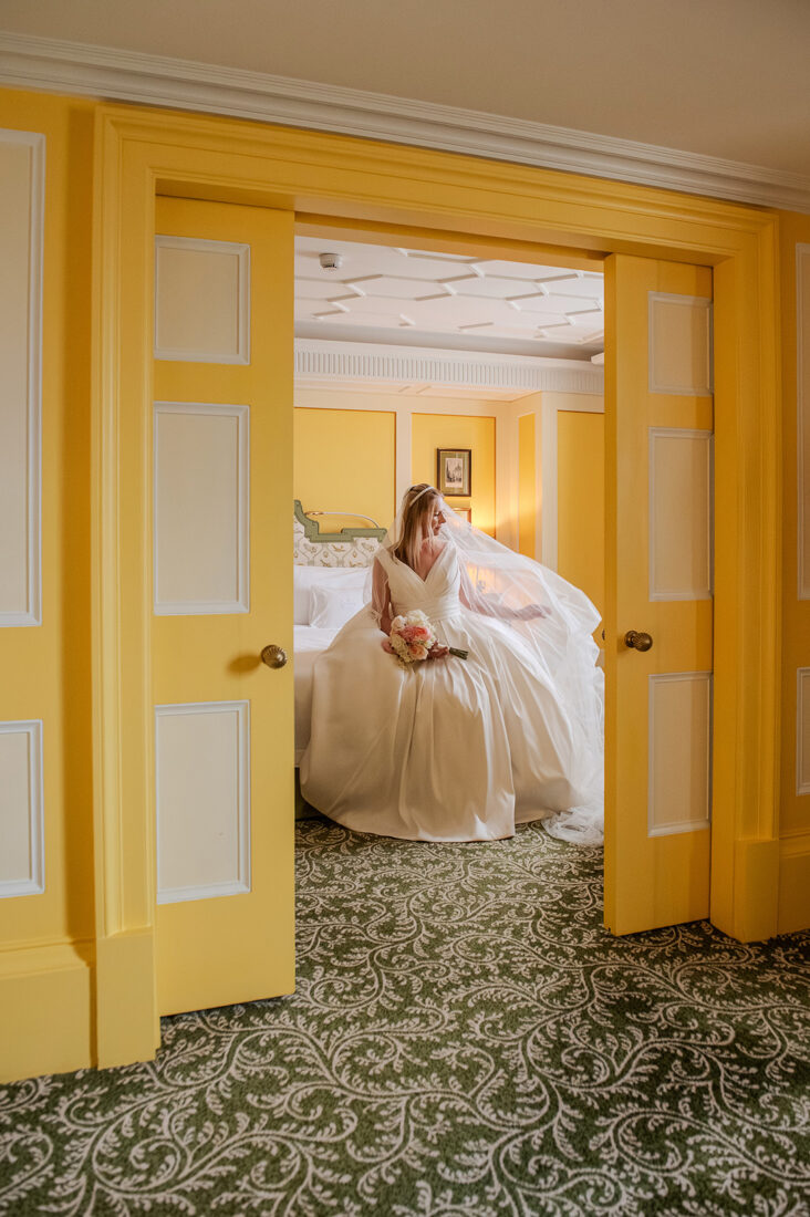 lanesbourough-bridal-suite-london-wedding - Wedding photographer London Bride in a white wedding gown holding a bouquet, standing in a doorway with yellow walls behind her and veil flowing.