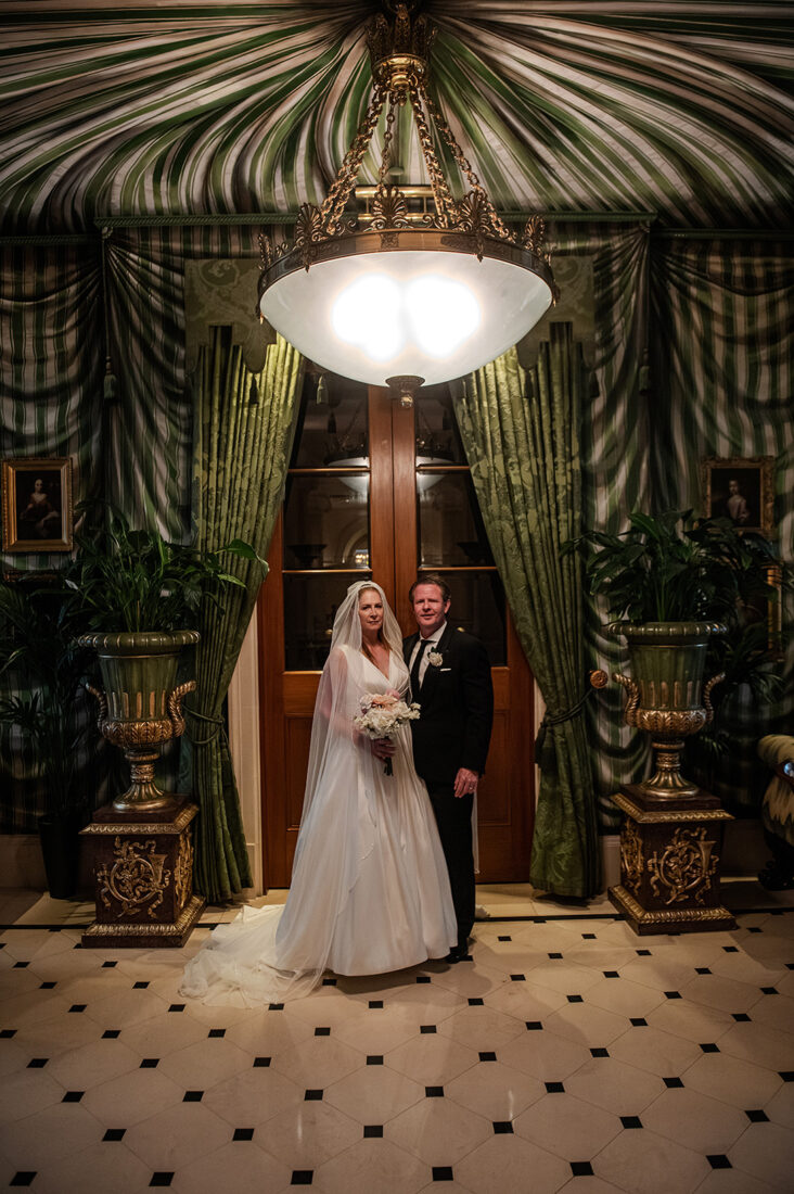 lanesbourough-hotel-photographer - Wedding photographer London Bride in a white wedding gown and veil holds a bouquet beside a groom in a black tuxedo under an ornate chandelier in a green-draped, formal foyer.