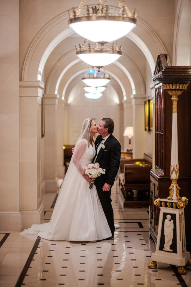 lanesbourough-hotel-wedding-couple - Wedding photographer London Bride in a white wedding gown kissing her groom in a marble hallway, of the Lanesborough holding a bouquet at her side