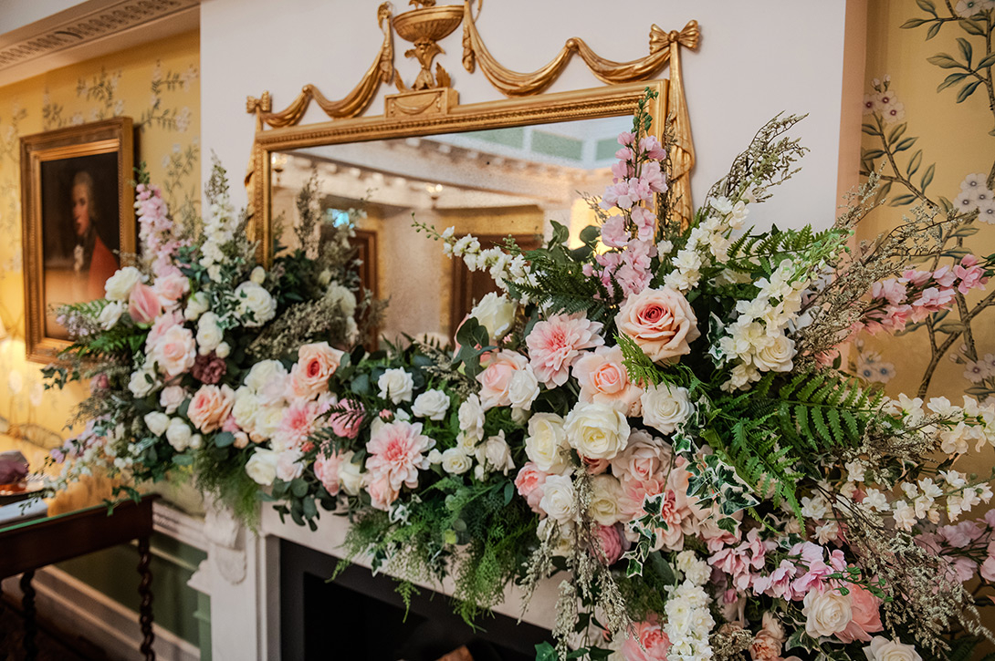 lanesbourough-wedding-flowers-photography - Wedding photographer London Elegant floral mantel arrangement with pink and white roses, peonies, and greenery on a marble fireplace, with a gilded mirror above.