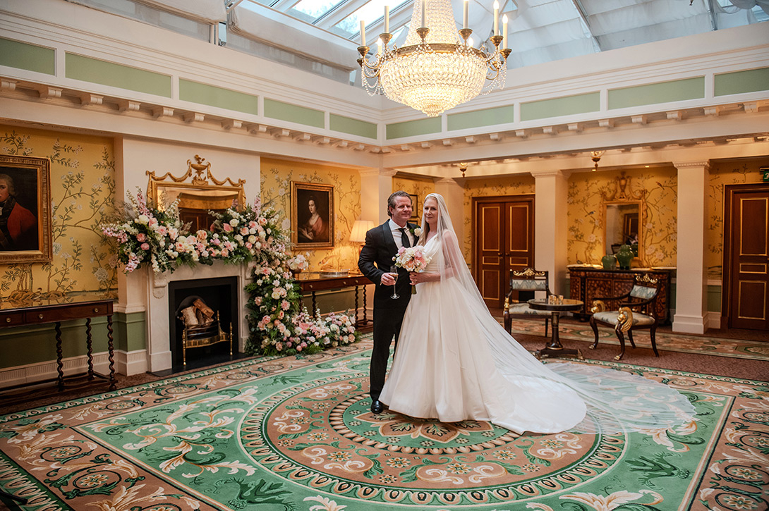 lanesbourough-wedding-photography-london - Wedding photographer London Bride and groom in formal attire pose with bouquet in an ornate, chandelier-lit ballroom.